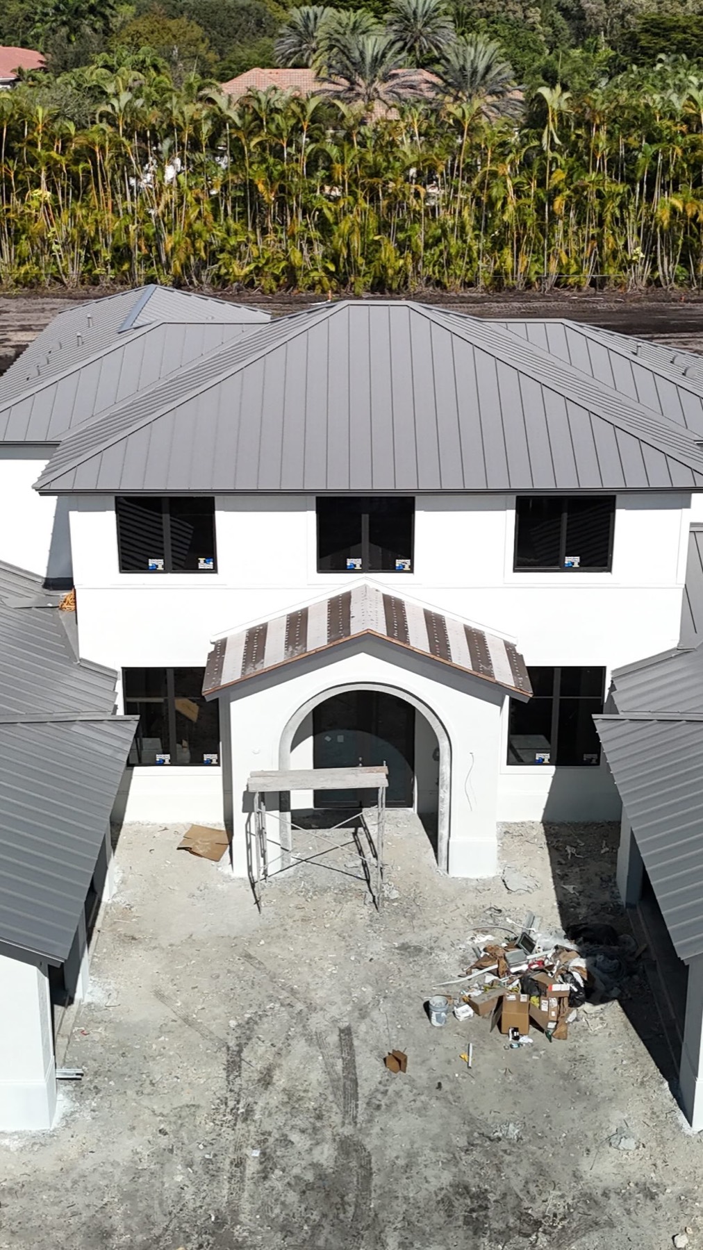 White Mediterranean home under construction with a slate-grey standing-seam metal roof, palms behind